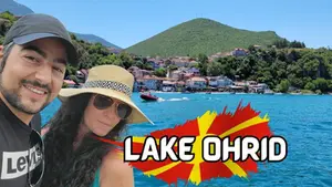 Smiling man and woman with hat by Lake Ohrid. Background: blue water, boat, green hills, houses. Text: Lake Ohrid. Sunny, vibrant scene.