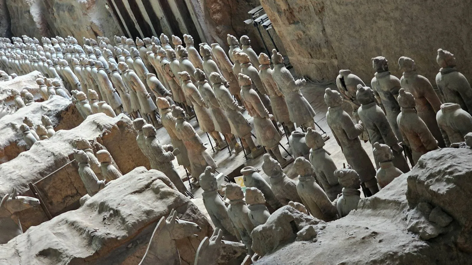 Rows of life-sized Terracotta Army statues stand partially unearthed in an archaeological excavation site, with some figures more exposed than others and earth walls surrounding them.