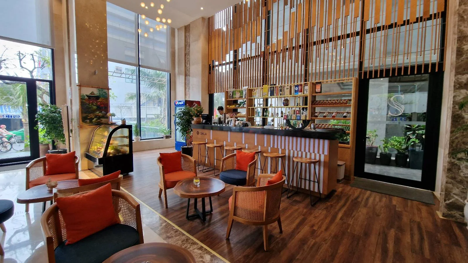 A modern café interior with wooden flooring, chairs, and round tables, featuring a bar counter with stools. Large windows allow natural light, and the decor includes plants and stylish hanging lights. Orange cushions add a pop of color.