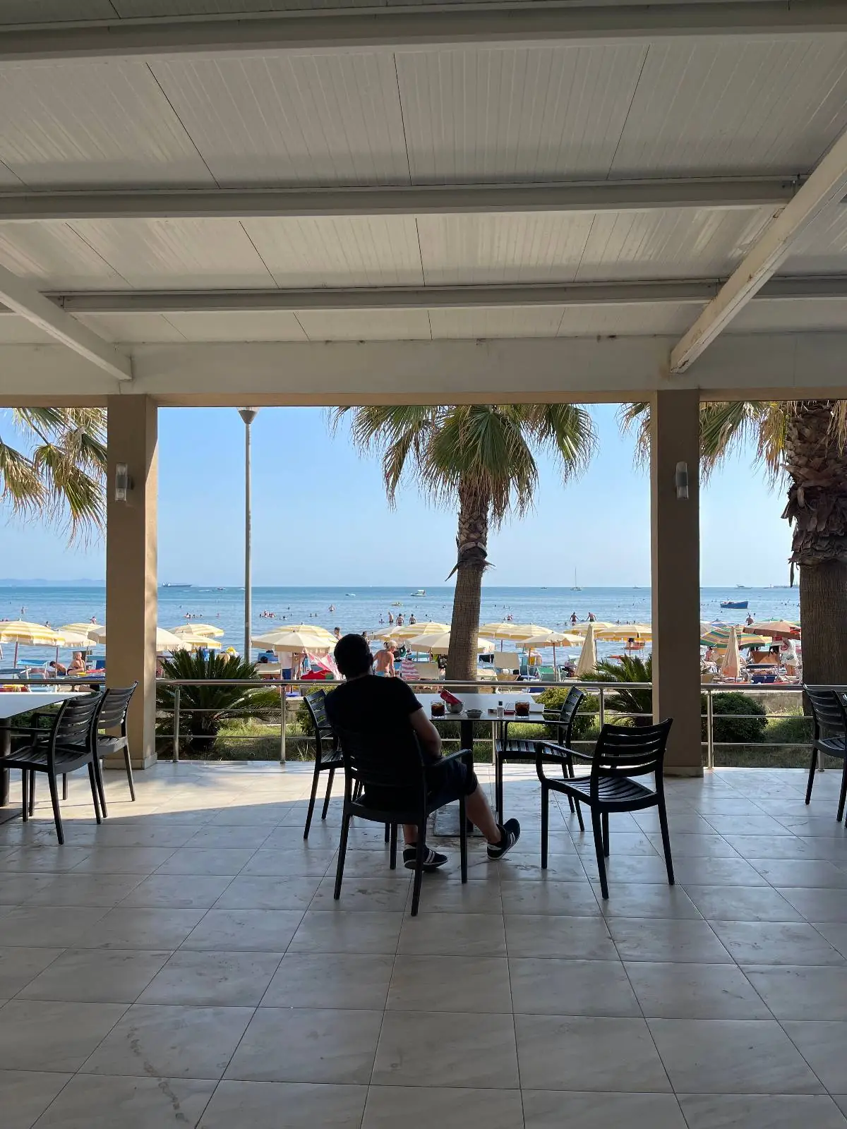 A person sits at a shaded patio table overlooking a beach with umbrellas, people, and palm trees on a sunny day. The ocean is visible in the background under a clear blue sky.