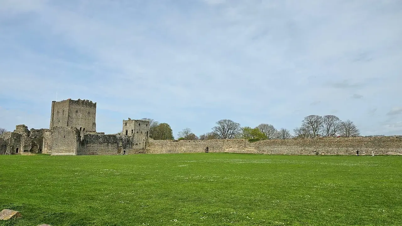 Ruins of a medieval castle with two prominent towers, surrounded by a large grassy field and a clear blue sky.