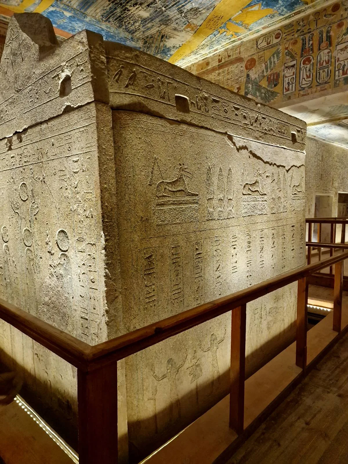 Ancient stone sarcophagus covered in intricate hieroglyphics, enclosed by a protective wooden railing. The ceiling above is colorful and ornate, suggesting a historical or museum setting.