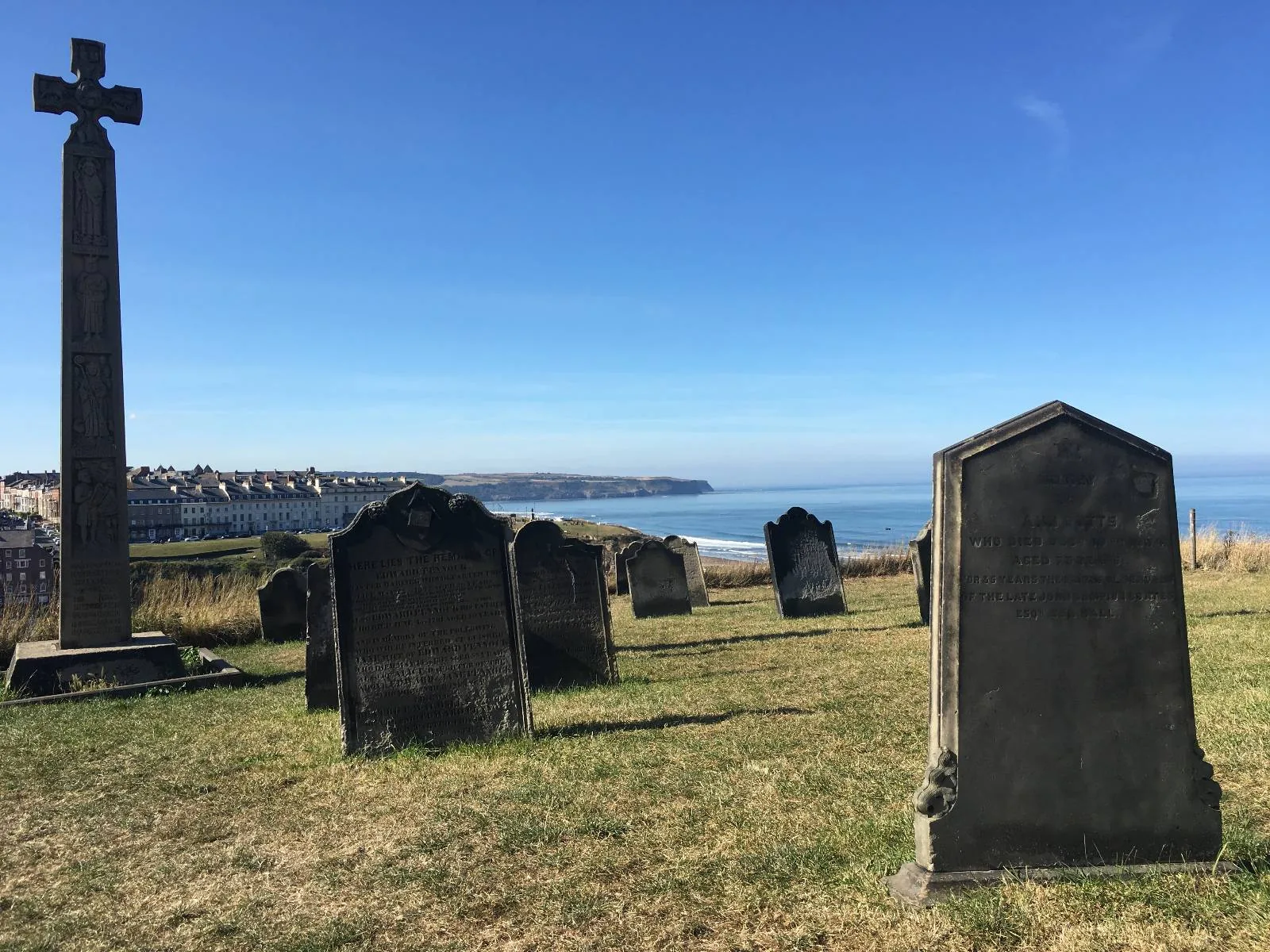 A graveyard with several tombstones and a tall cross-shaped monument under a clear blue sky. In the background, there is an ocean view and a coastline.