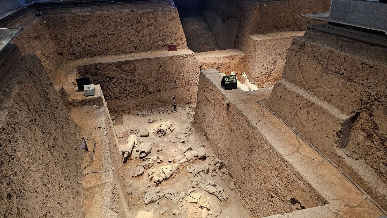 Large archaeological excavation site with deep, stepped earth walls and scattered artifacts in the bottom, possibly bones or relics, illuminated by overhead lights.