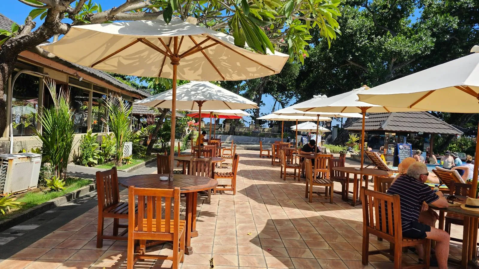 Outdoor restaurant patio with wooden tables and chairs under large white umbrellas. People are seated, enjoying food and drink, surrounded by greenery and trees on a sunny day.