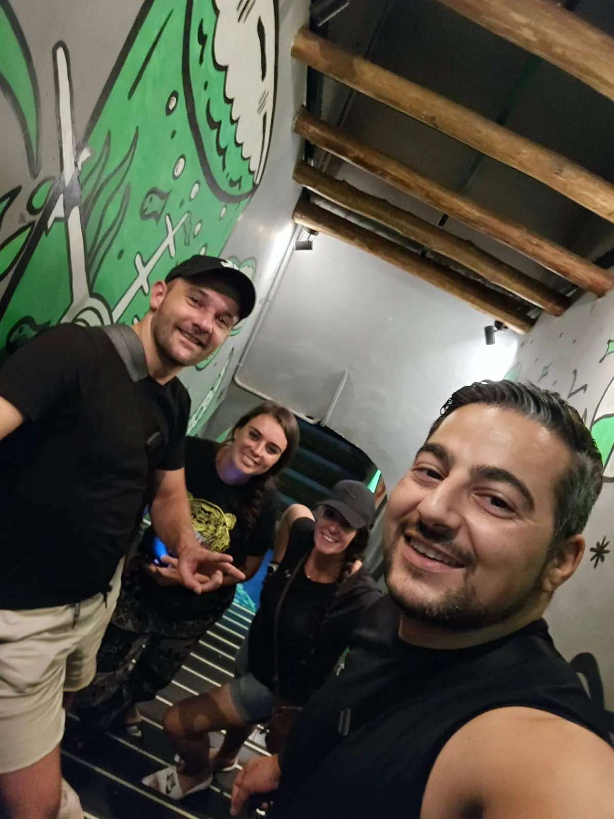 Four people smiling on a decorated staircase with green wall art. Two are on the left, one wearing a hat, the other in a black shirt and shorts. On the right, two women, one with long hair and another wearing a cap, all appear happy and relaxed.
