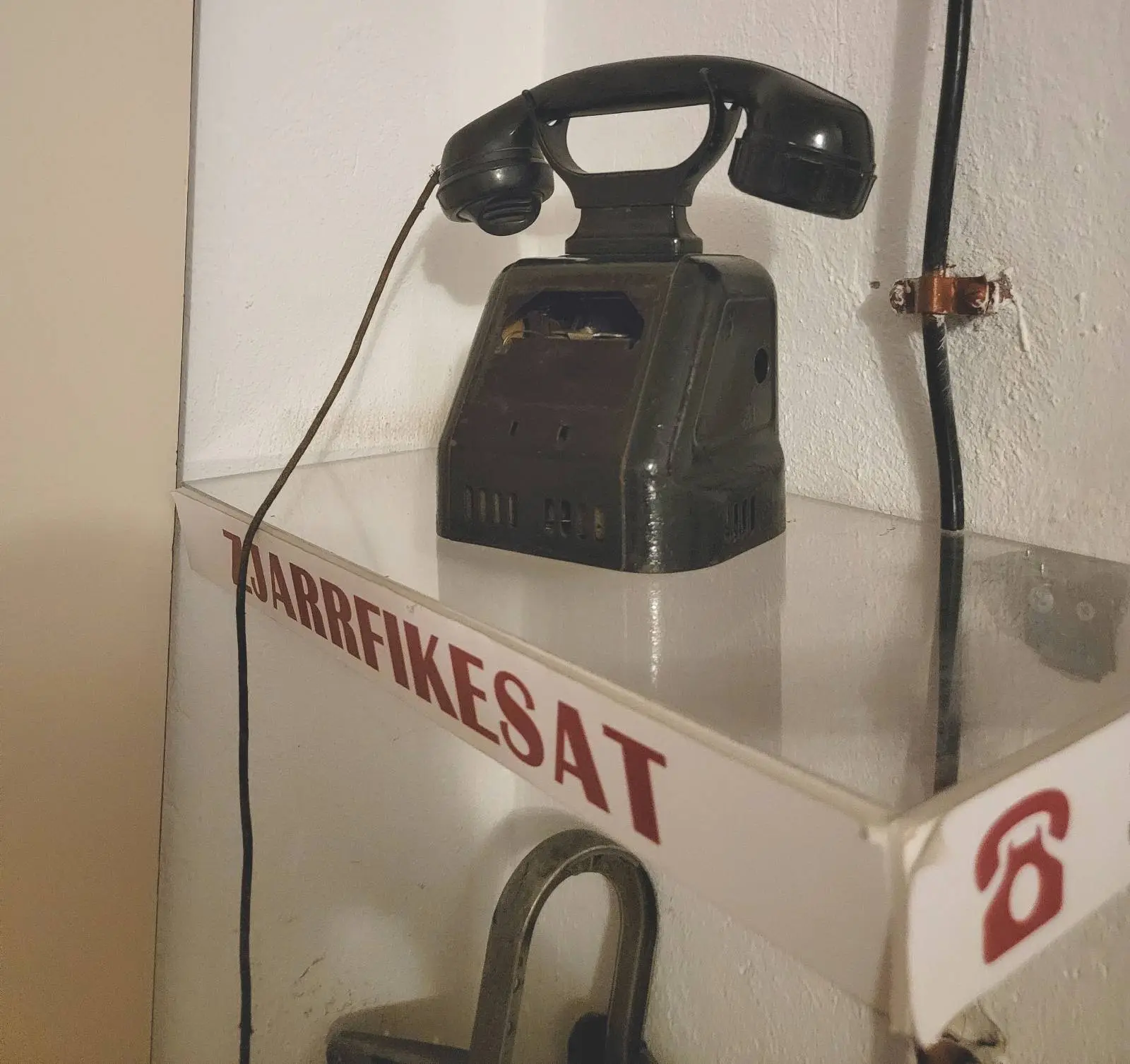 A vintage black rotary telephone sits atop a white metal box with the text "TARIFIKESAT" on it. A red telephone symbol is visible on the box. The phone and box are placed against a white wall.