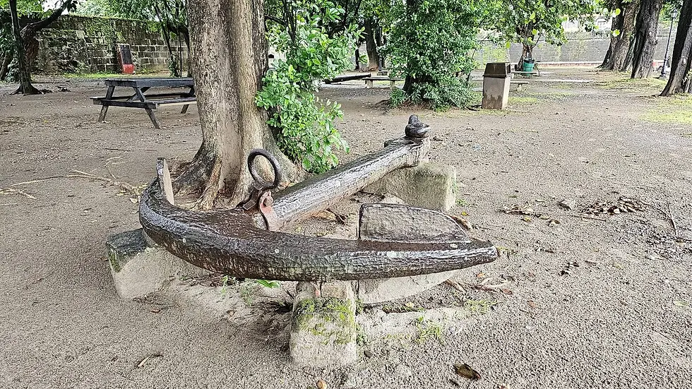 Rusty anchor rests against a tree in a park with benches and greenery, creating a serene atmosphere. Overcast sky visible through trees.