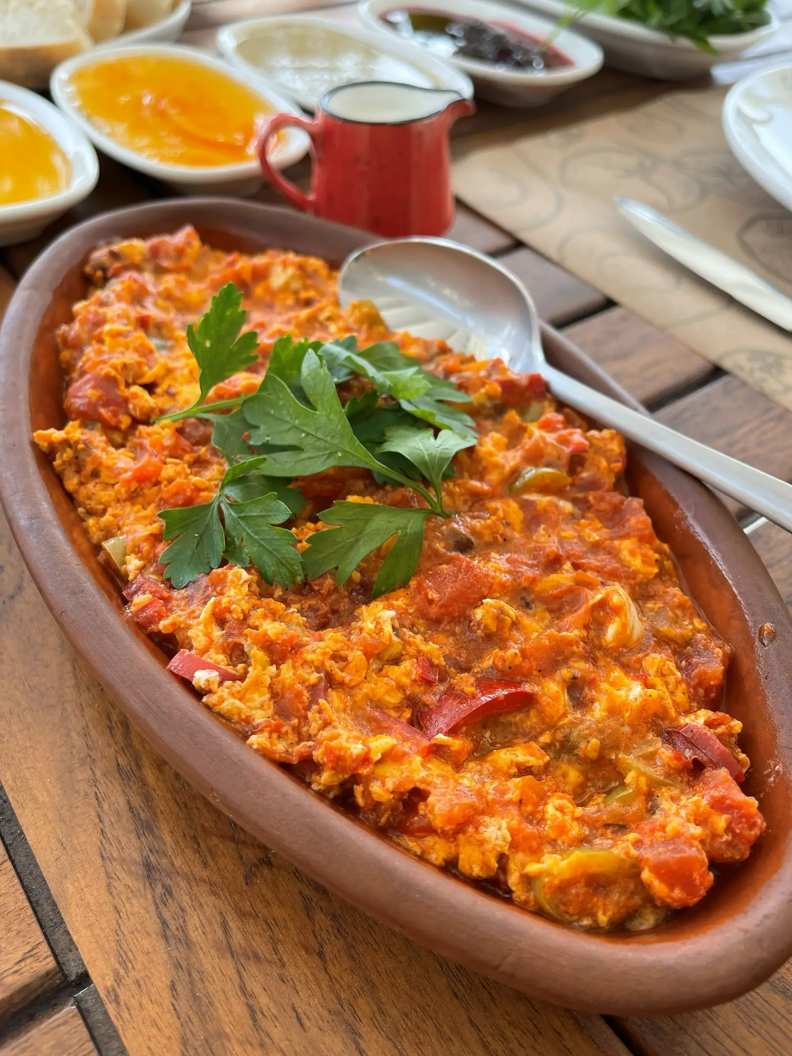A dish of scrambled eggs with tomatoes and herbs is served in a brown oval dish, garnished with parsley, with a spoon on the side. Various condiments are visible in the background.