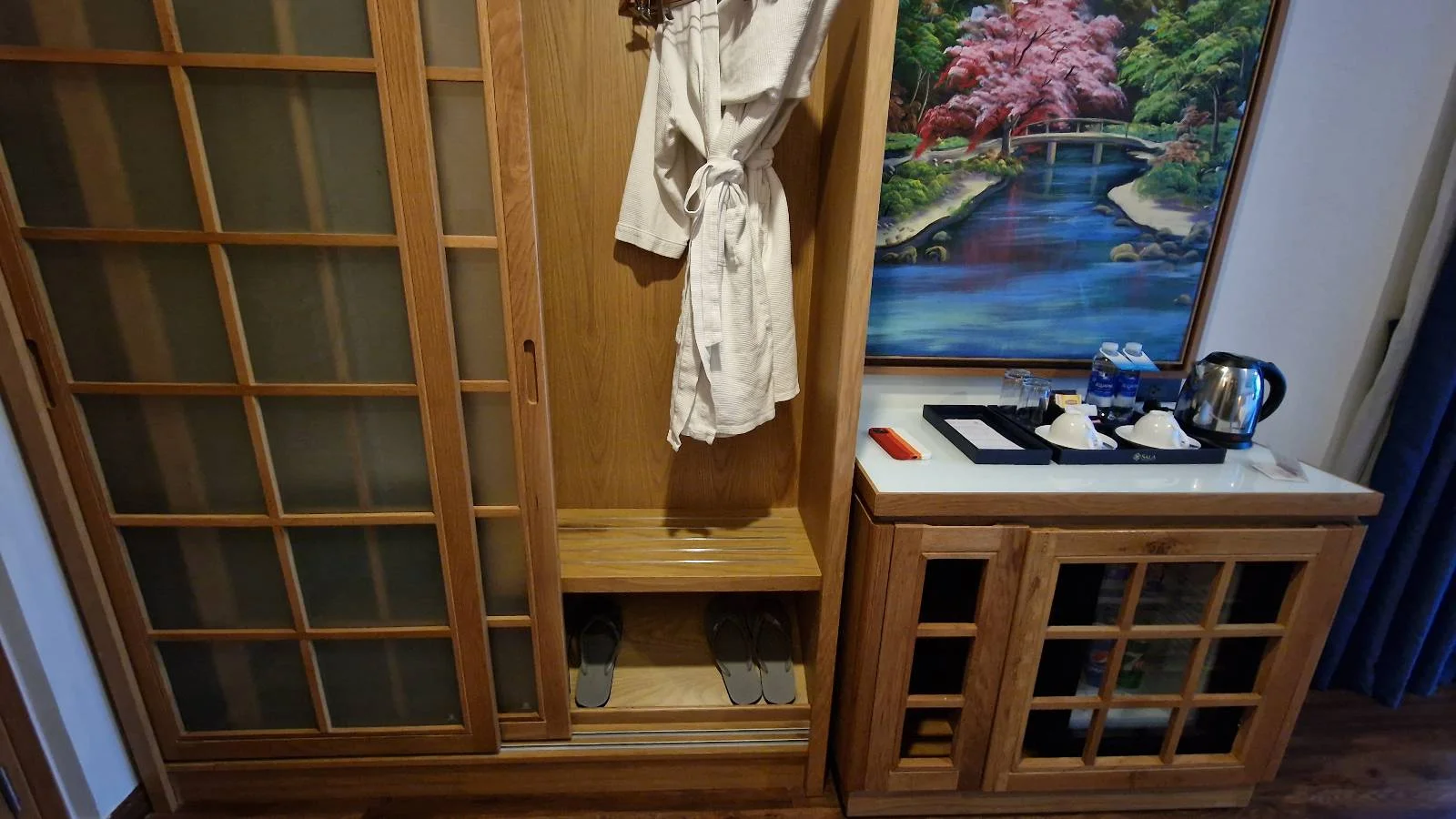 A wooden wardrobe with an open sliding door reveals a hanging robe. Next to it is a wooden cabinet with a tray holding a kettle and cups. A framed painting of a serene garden scene hangs above the cabinet.