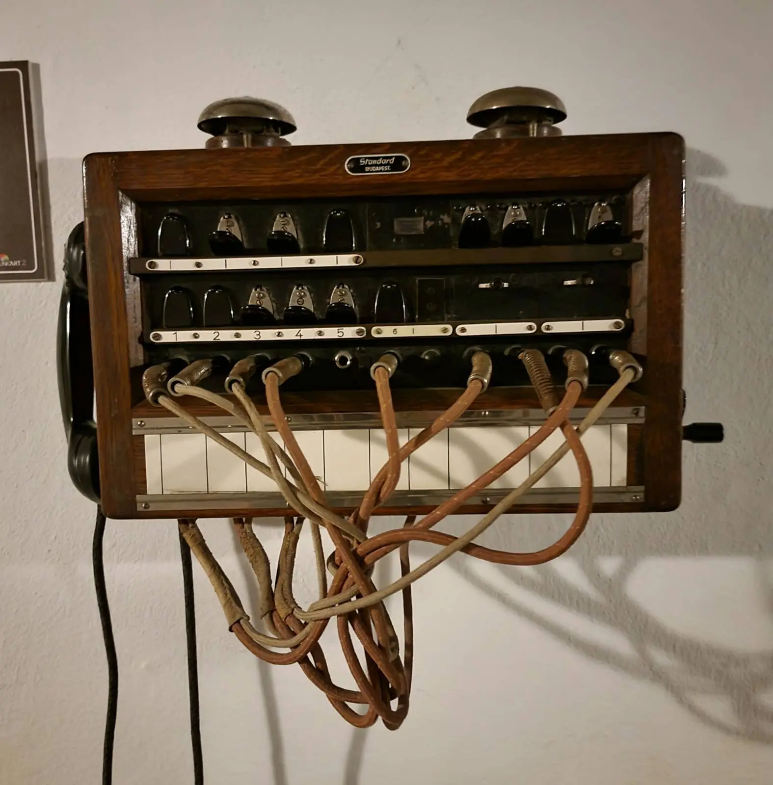 A vintage telephone switchboard with multiple cables and switches, mounted on a wall. The device is enclosed in a wooden frame, showing a tangle of wires connected to the lower part. The setup appears old and utilitarian.