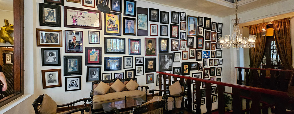 A cozy room with a wall covered in various framed photos and artworks. The space features a seating area with beige cushions and a wooden table, surrounded by chairs. Warm lighting comes from an elegant chandelier hanging above.