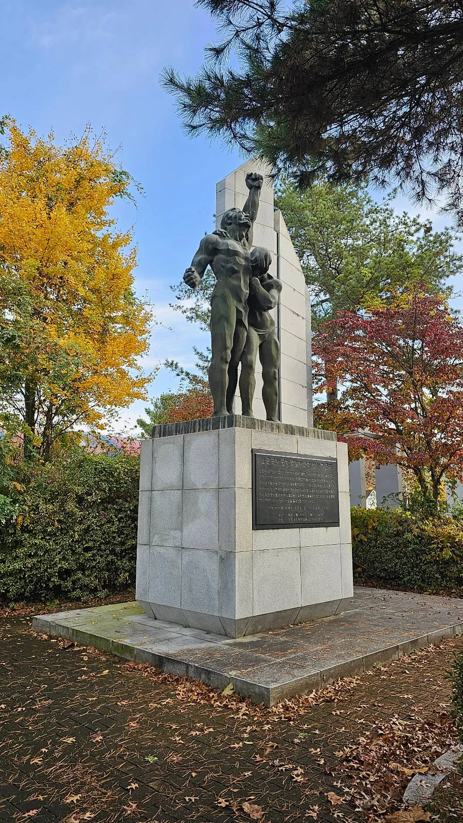 A tall stone monument with a bronze statue of two figures holding a torch, set in a leafy park. The trees surrounding the monument display vibrant autumn colors of yellow, orange, and red. The ground is covered with fallen leaves.