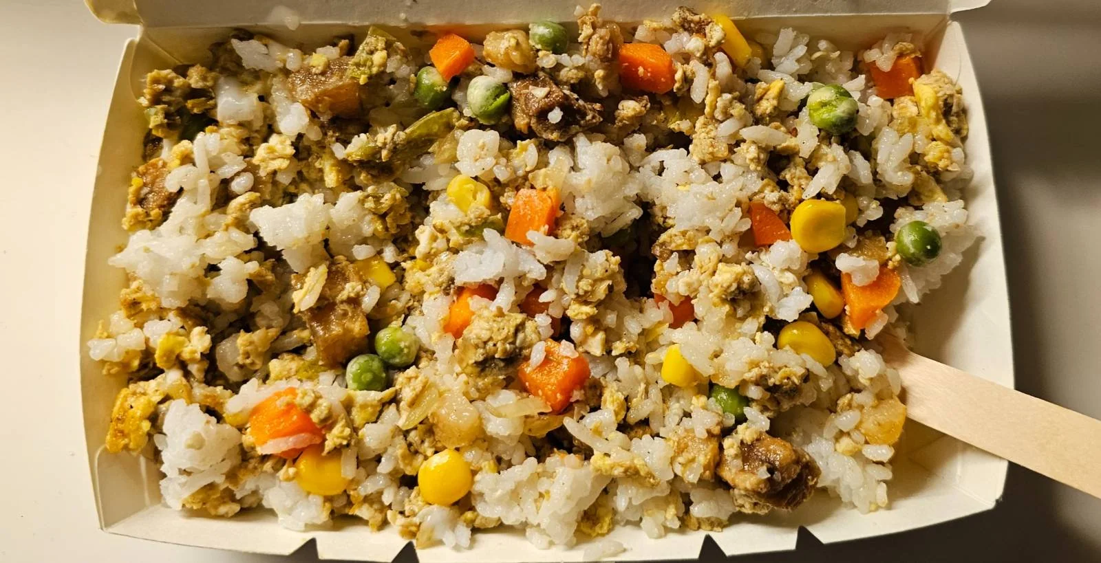A takeout container filled with fried rice mixed with vegetables such as carrots, peas, and corn, along with pieces of scrambled egg and possibly meat. A wooden utensil is visible on the right side.