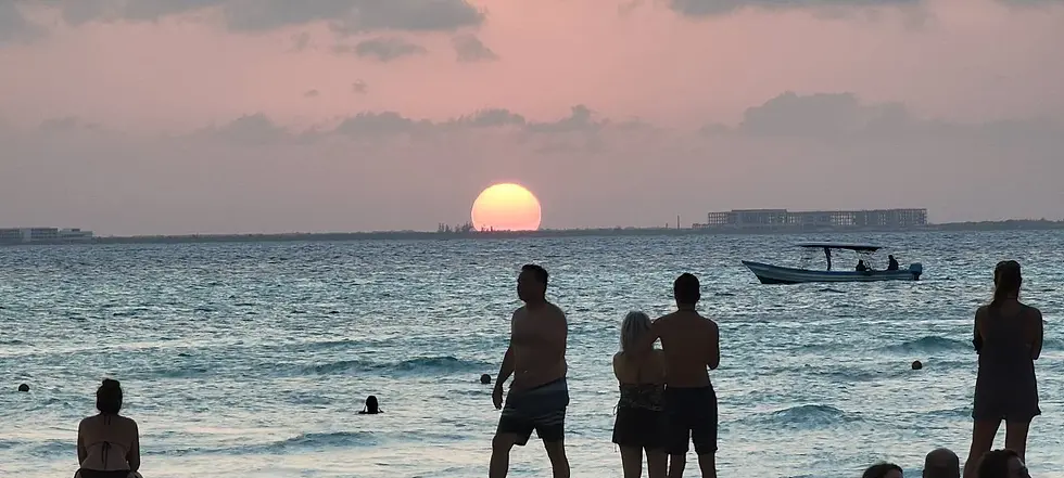 burnt orange sun setting on the horizon of blue water, with several people swimming. Three people stand and watch from the beach and there is also a white sailboat in the distance