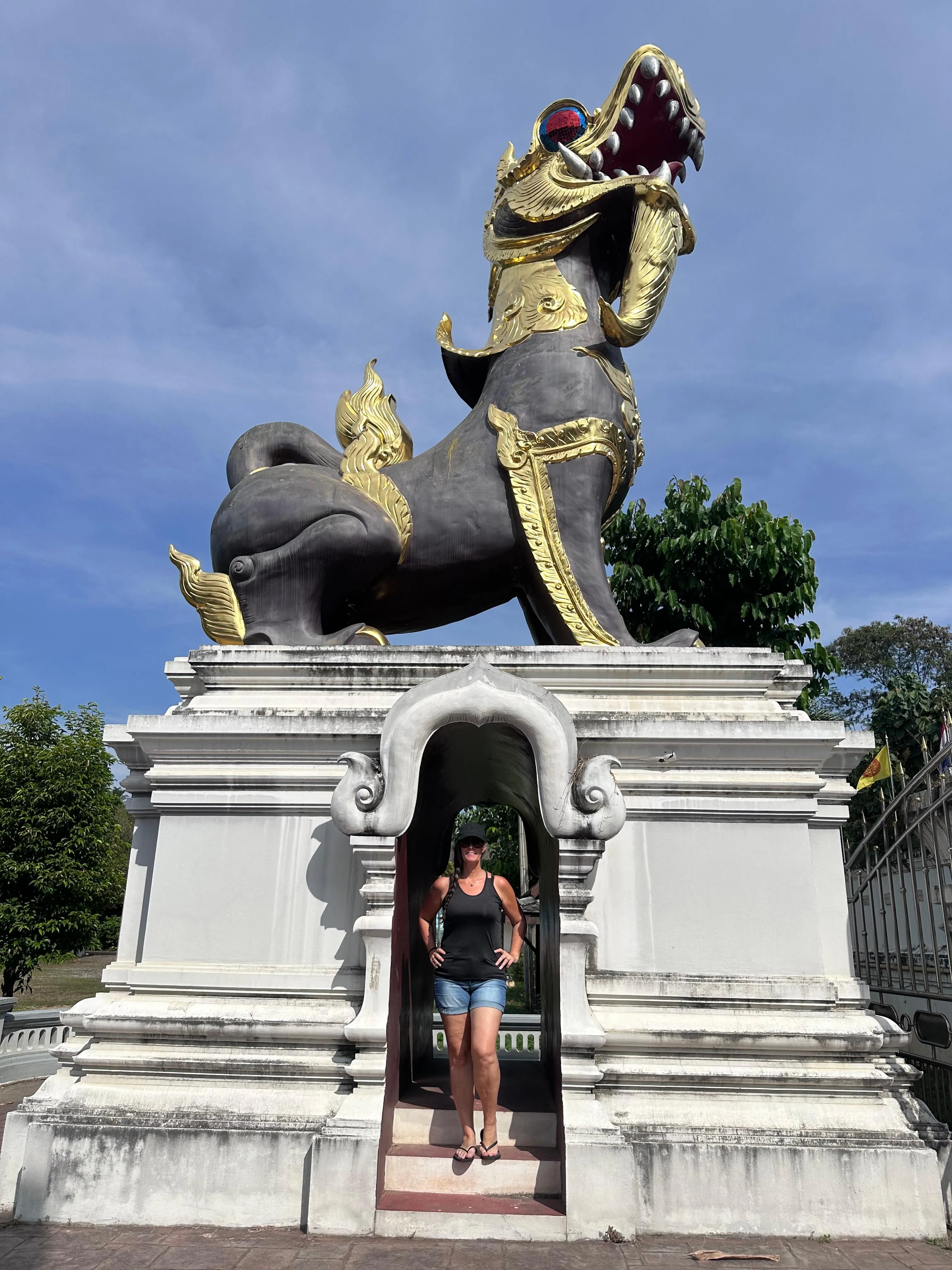 A person stands beneath a large, ornate statue of a mythical lion-like creature with gold decorations, set on a white pedestal outdoors under a blue sky.