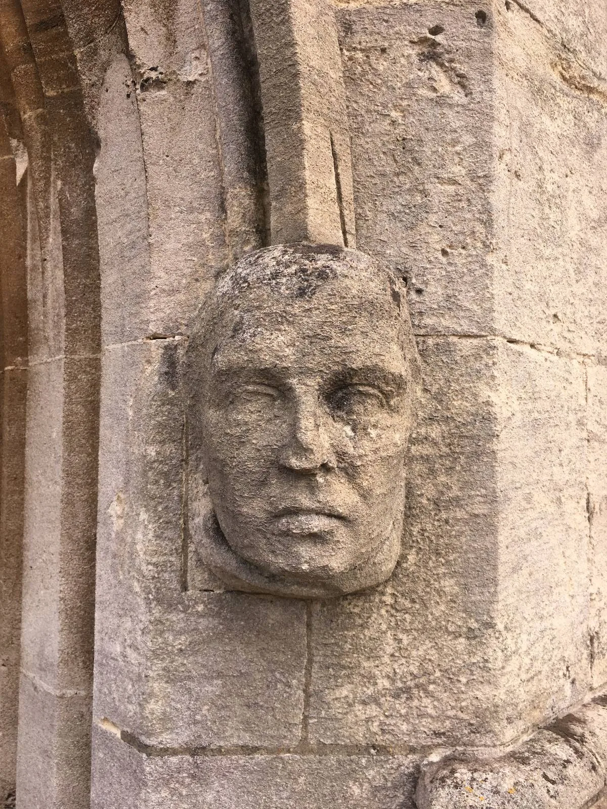 Stone carving of a human head embedded in a weathered stone wall, featuring a solemn expression. The carving is aged, suggesting historical significance, and is lit by natural light, highlighting its intricate details.