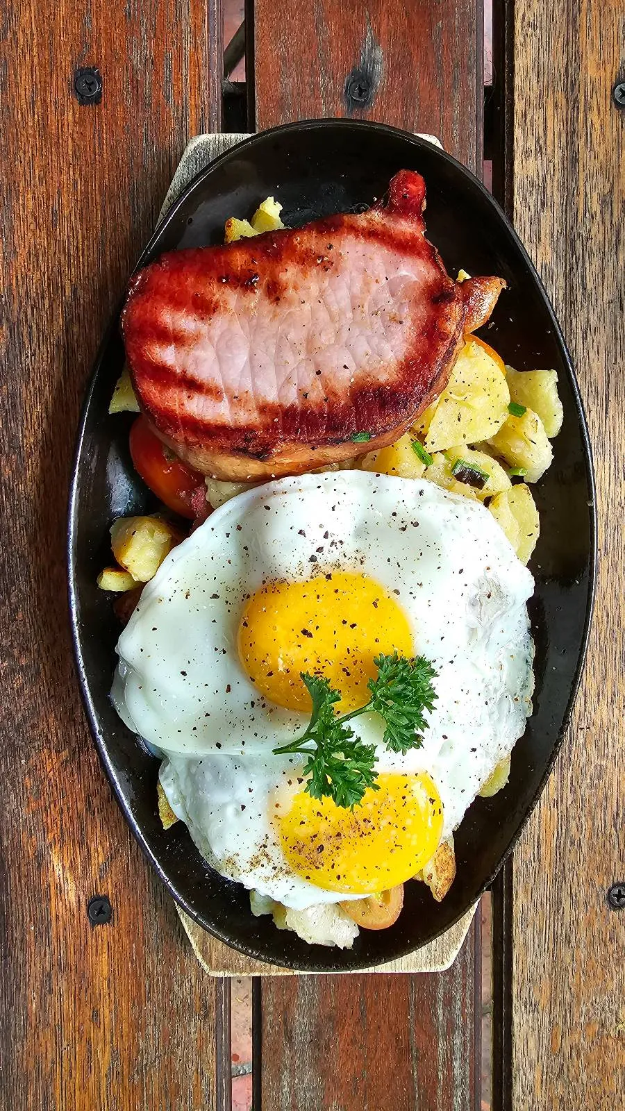 A grilled slice of meat sits on a bed of roasted potatoes and vegetables, topped with two sunny-side-up eggs garnished with fresh parsley, all served on a dark oval plate placed on a wooden table.