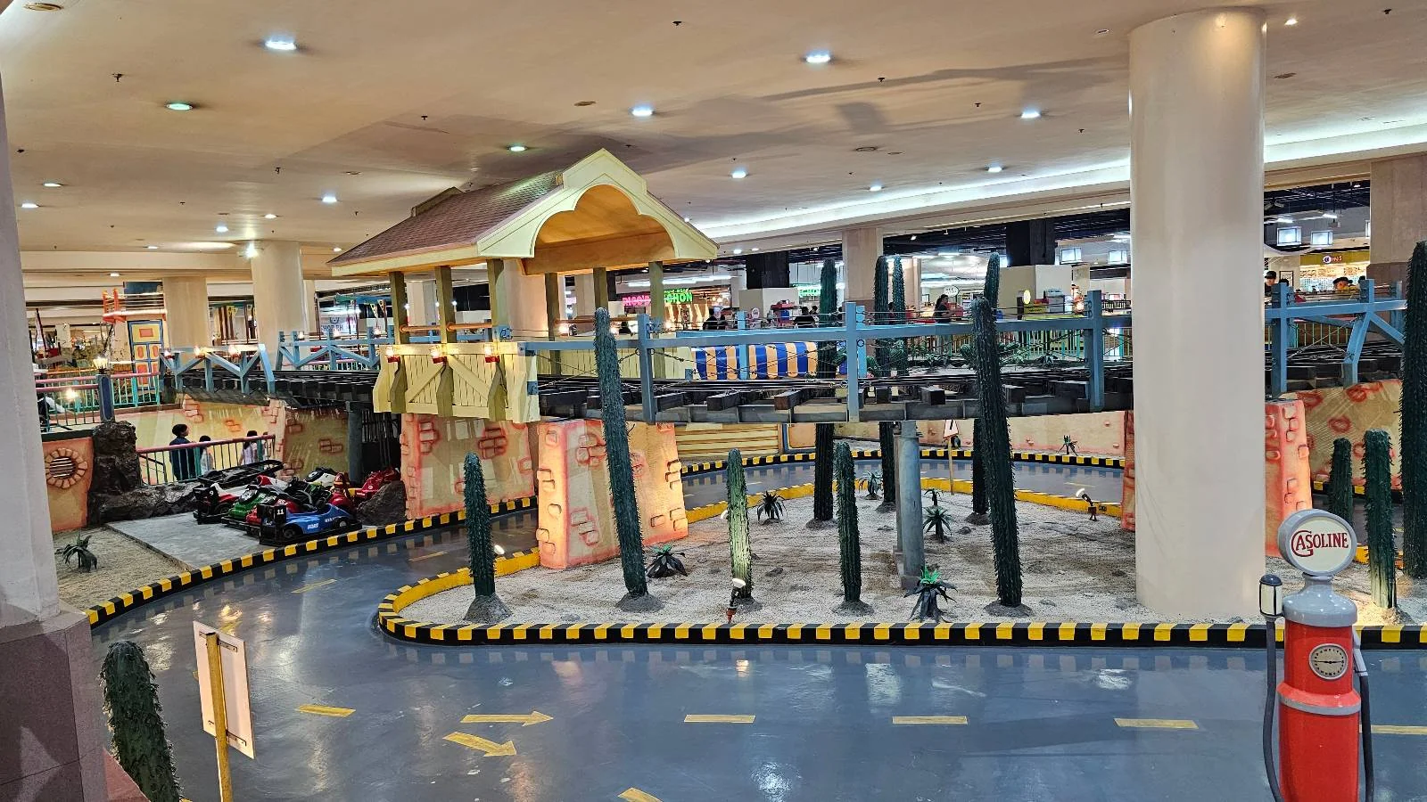 Indoor go-kart track with racing cars and a western-themed decor, featuring artificial structures resembling wooden buildings and cacti. The track is surrounded by shops and lit by ceiling lights in a spacious area.