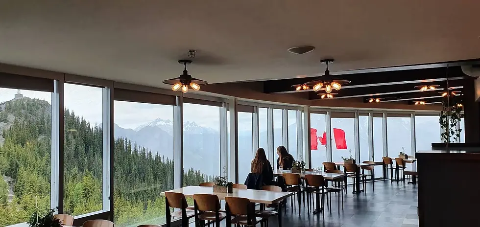 Interior of a restaurant with large windows offering a view of a forested mountain landscape. Two people are seated, and a Canadian flag is visible outside.