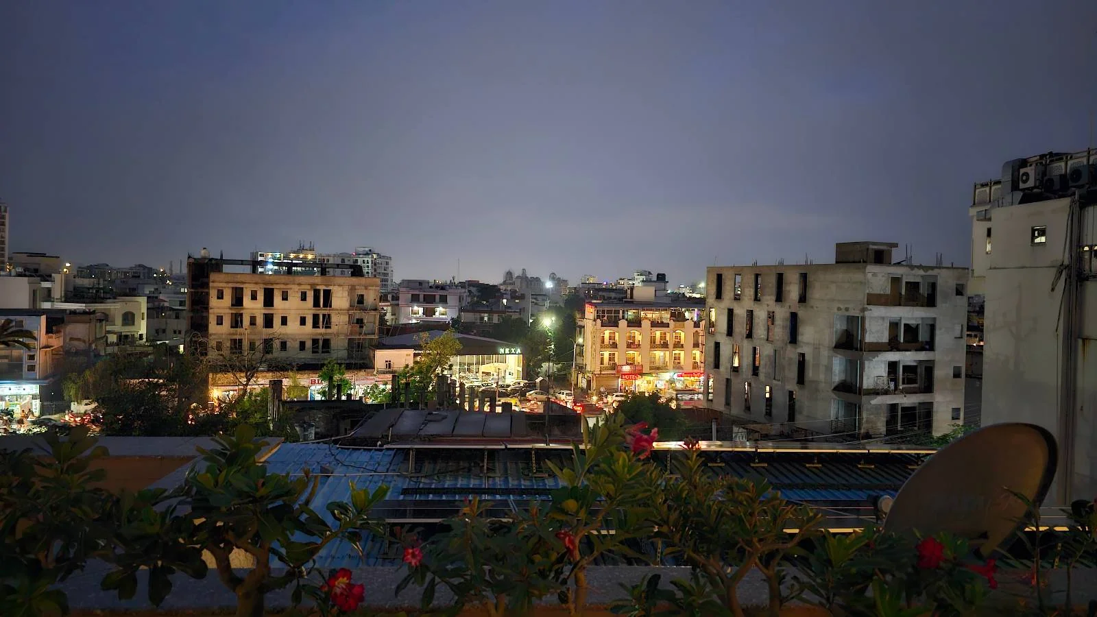 Cityscape at dusk showing several apartment buildings with lights on, a few trees and plants in the foreground, and a cloudy sky above. The scene appears calm with little activity visible.
