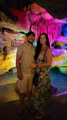 In a vibrant cave within Mexico's cenotes, a couple stands mesmerized by the illuminated rock formations. The man in a cap and shorts and the woman in a floral dress are enchanted by hues of pink, purple, yellow, and green - a scene reminiscent of Playa del Carmen's beauty.