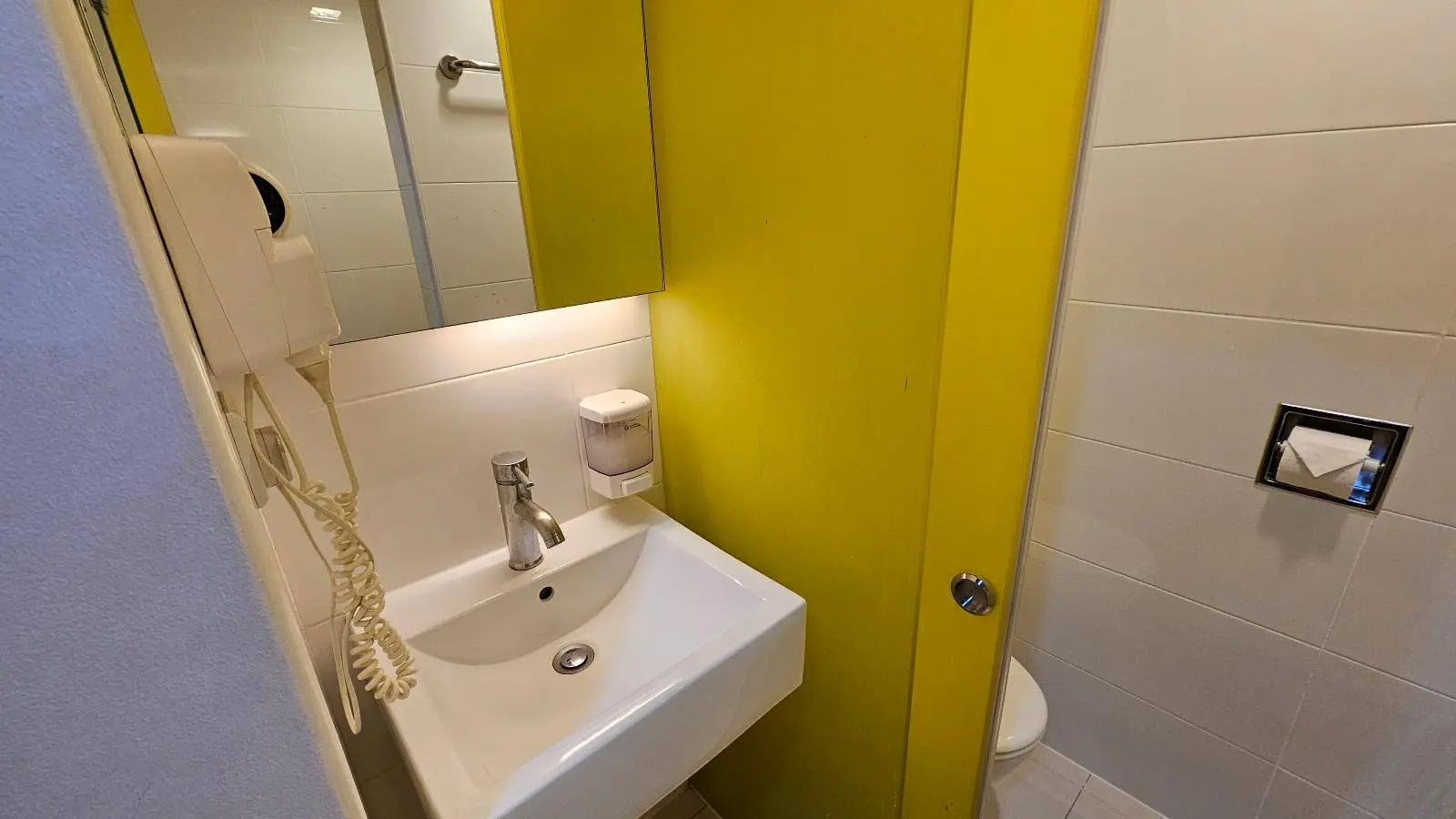 A small bathroom with a white sink, mirror, soap dispenser, telephone, and a partially visible toilet separated by a bright yellow partition. White tiled walls add to the clean, modern look.