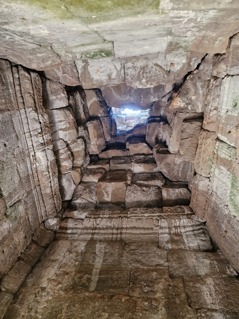 View looking up through a narrow, stone-lined shaft or tunnel with rough, stacked rocks, leading to a small opening at the top where daylight is visible.