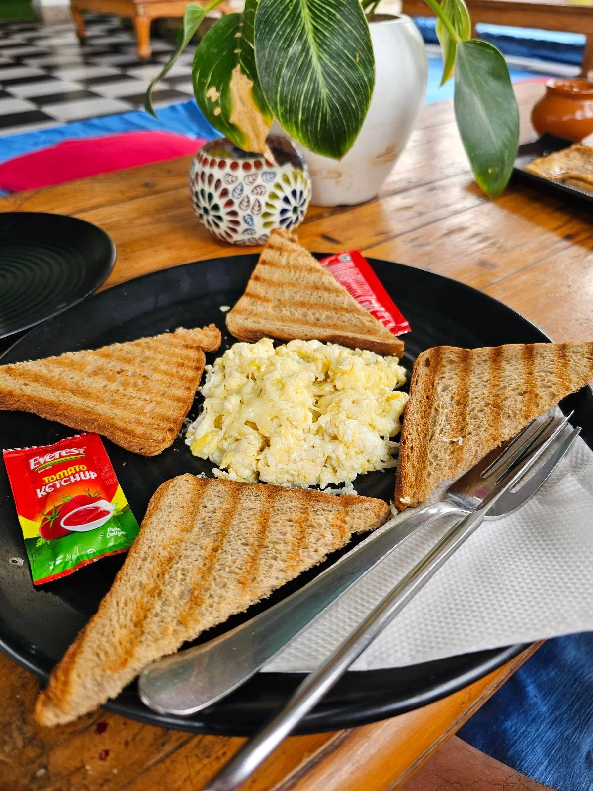 A black plate with four triangular toasted bread slices, a serving of scrambled eggs in the center, two small packets of butter and jam, a knife and fork, on a wooden table with a potted plant in the background.