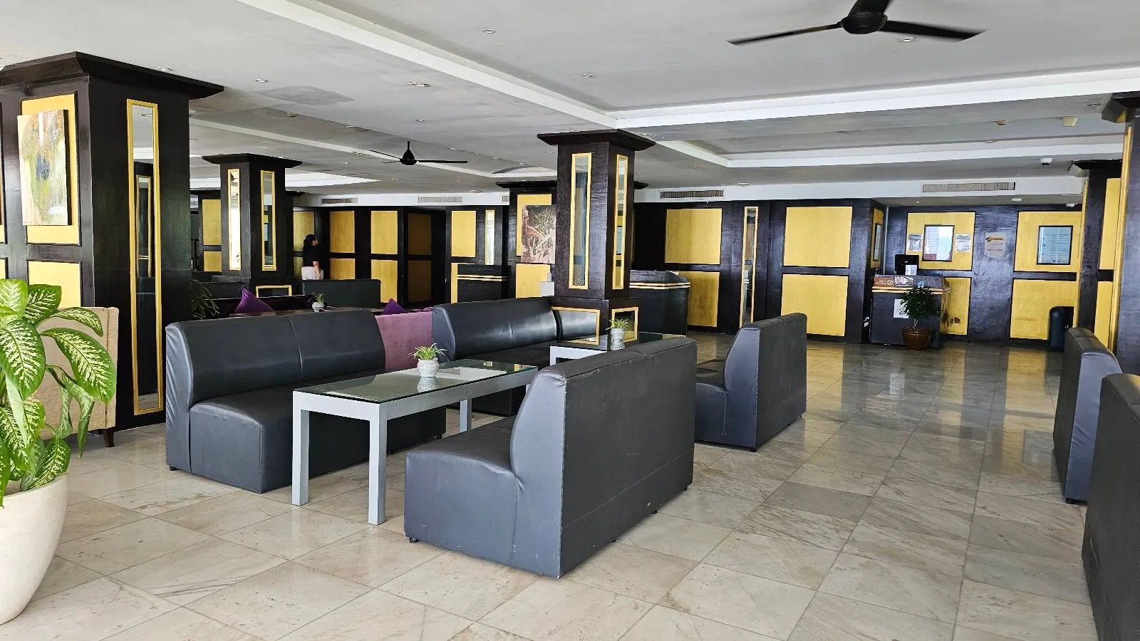 A spacious hotel lobby with polished floors, modern black leather seating, and glass-topped coffee tables. Large potted plants and contemporary décor accentuate the area, while ceiling fans provide ventilation. A reception desk is visible in the background.