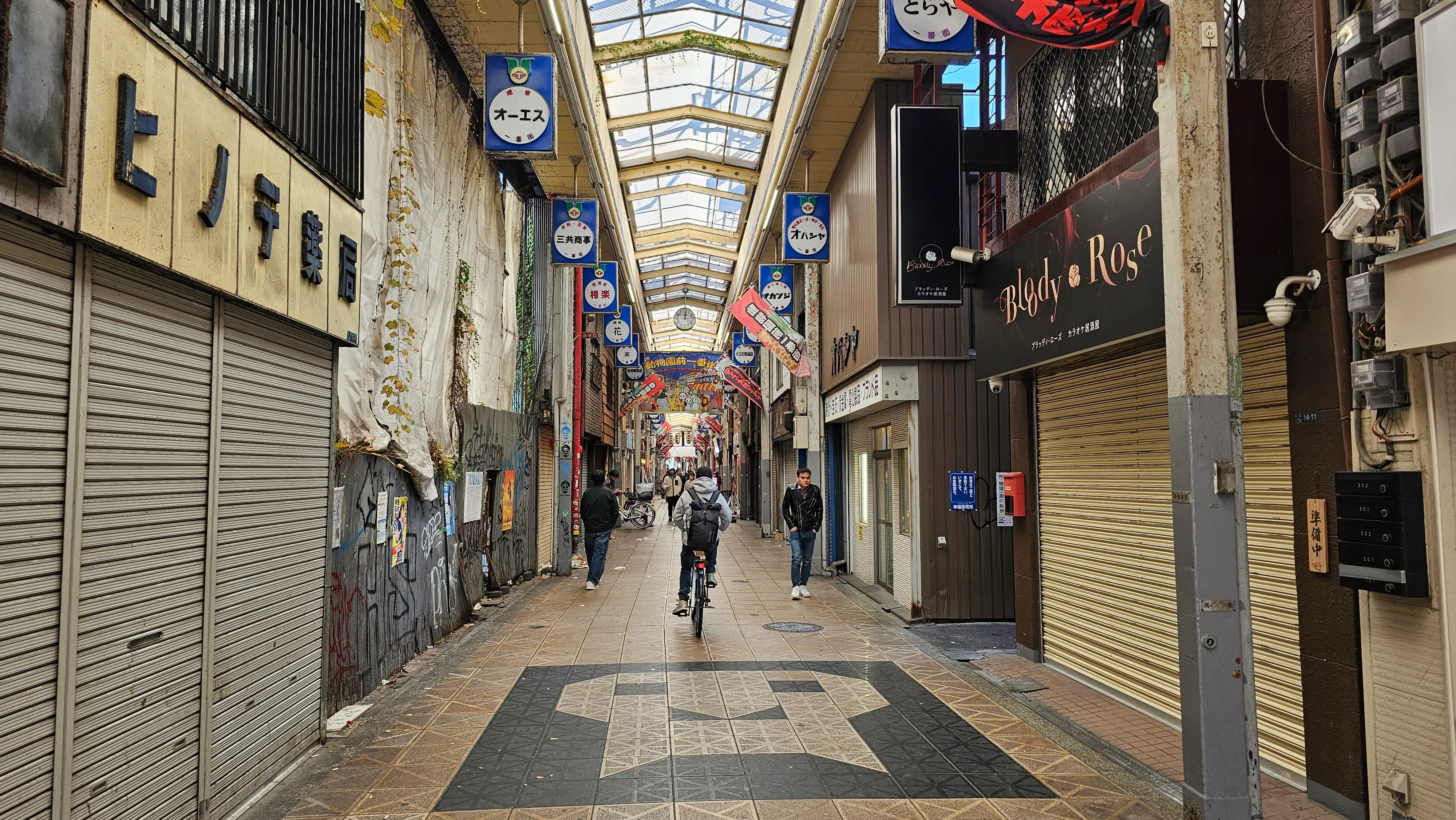 A covered shopping street in Japan with closed shop shutters, a few people walking, and a cyclist riding down the center. Colorful signs and banners hang from the ceiling, and natural light enters through the glass roof.