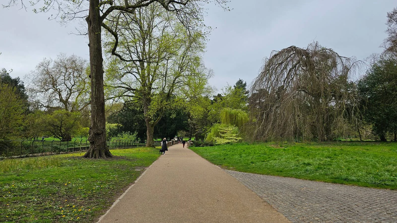 A paved path in a lush park with people walking, surrounded by green trees and a large rock formation on the right.