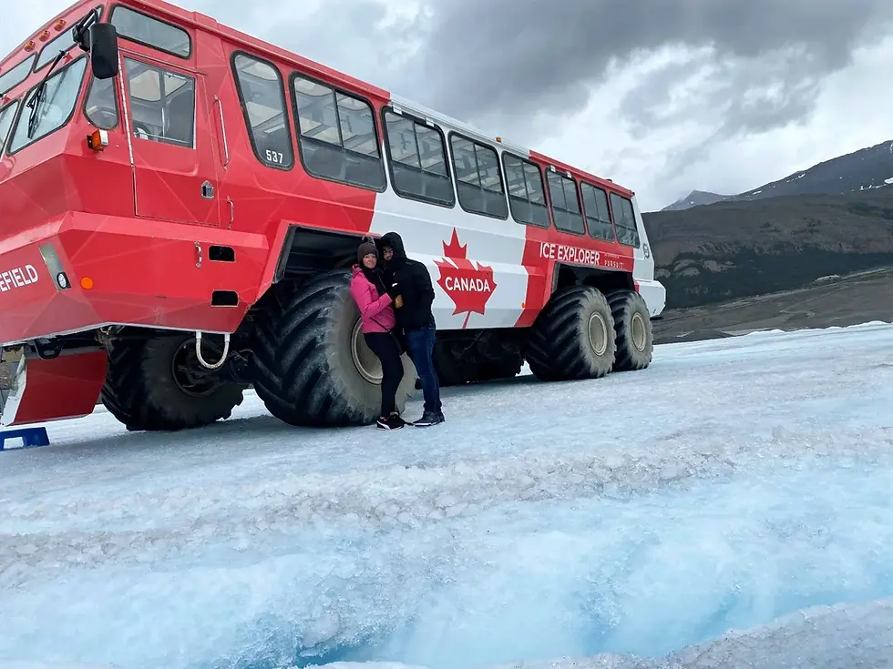 Couple poses by a large red and white Ice Explorer vehicle with "Canada" text, on a glacier. Cloudy sky and distant mountains in the background.