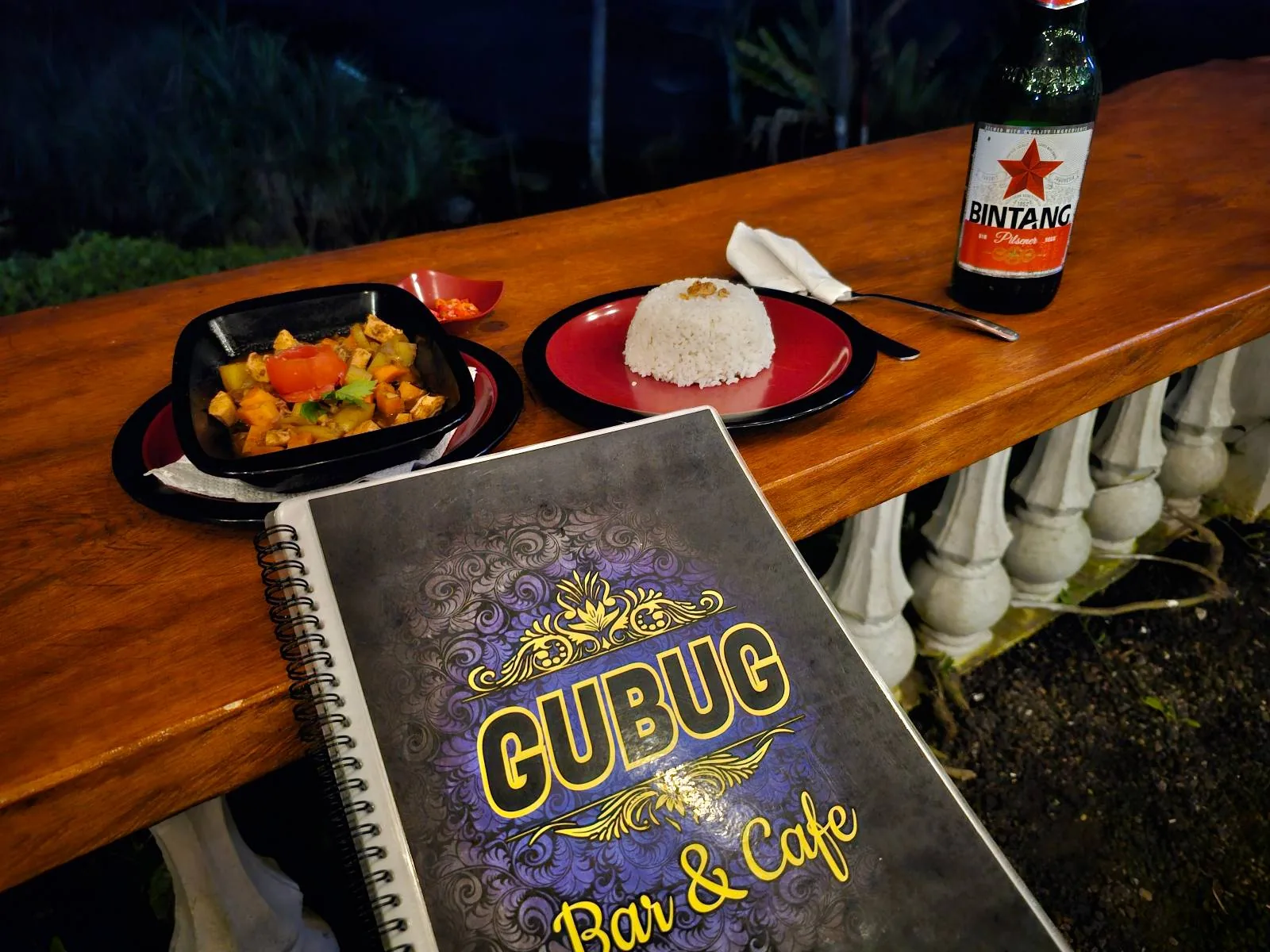 A wooden table holds a plate of food with vegetables and meat, a mound of white rice, and a cold beer bottle. A menu labeled "Cubug Bar & Cafe" is in the foreground. The background shows a nighttime outdoor setting.