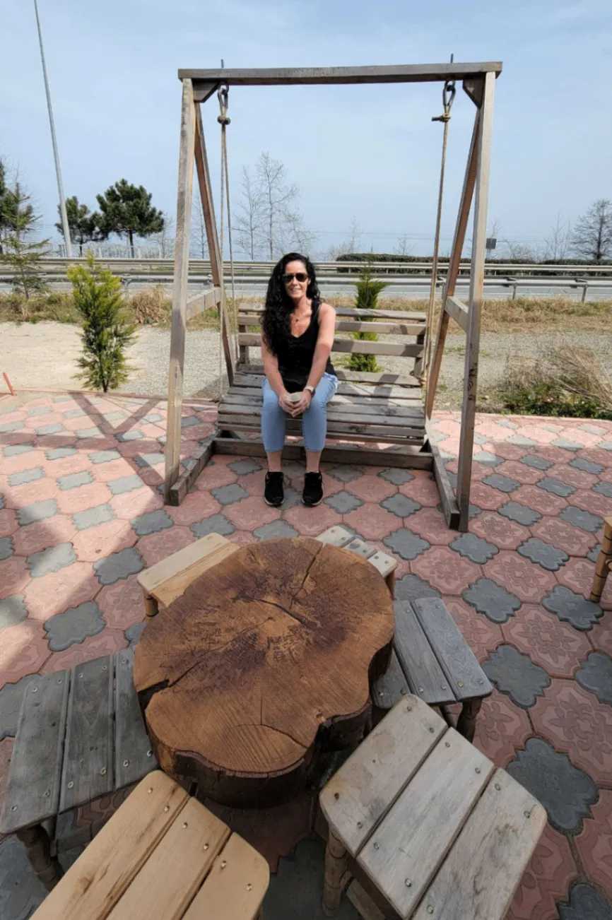 A person sits on a wooden swing in an outdoor area with a circular wooden table and benches on a patterned stone patio. Trees and a road are visible in the background.