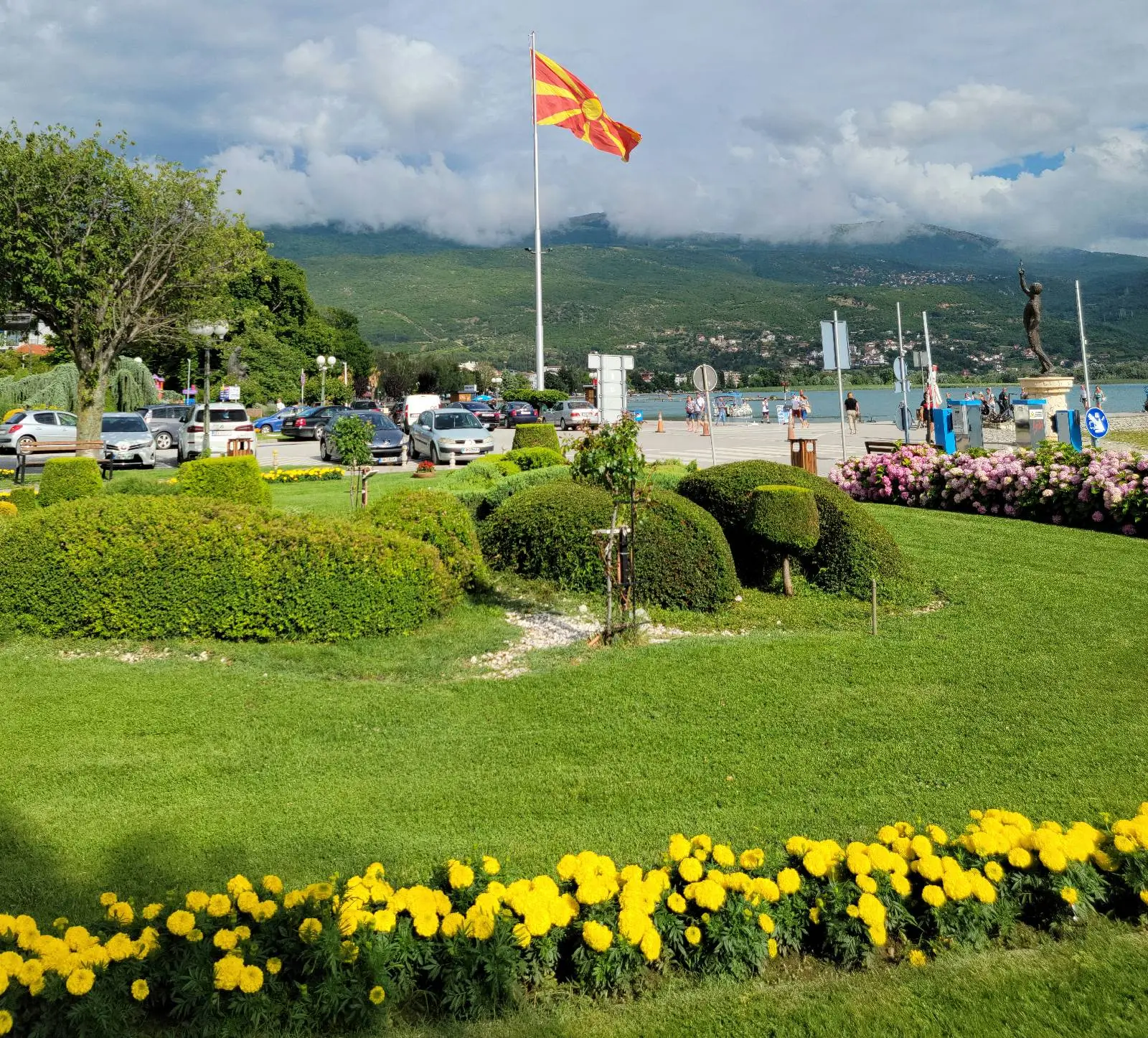 A landscaped park with neatly trimmed bushes and vibrant yellow flowers in the foreground. A Macedonian flag flies on a tall pole by a lakeside, with hills and a cloudy sky in the background. Cars and people are visible near the water.