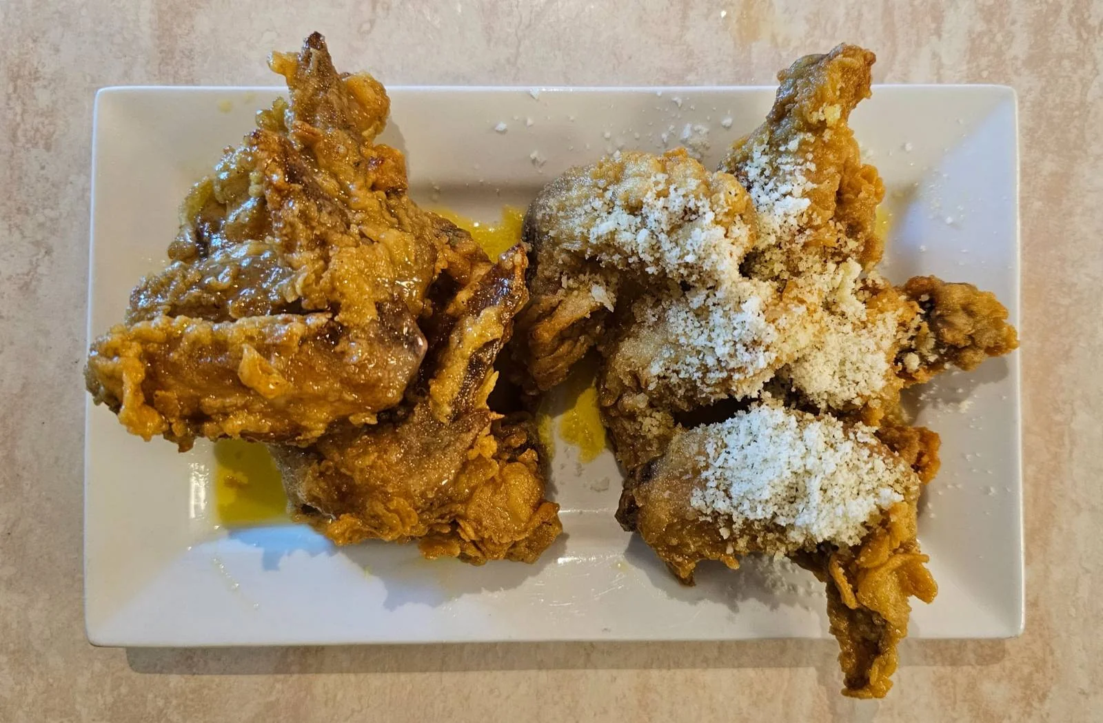 Two pieces of fried chicken on a rectangular white plate, sprinkled with grated cheese and placed on a light-colored surface.
