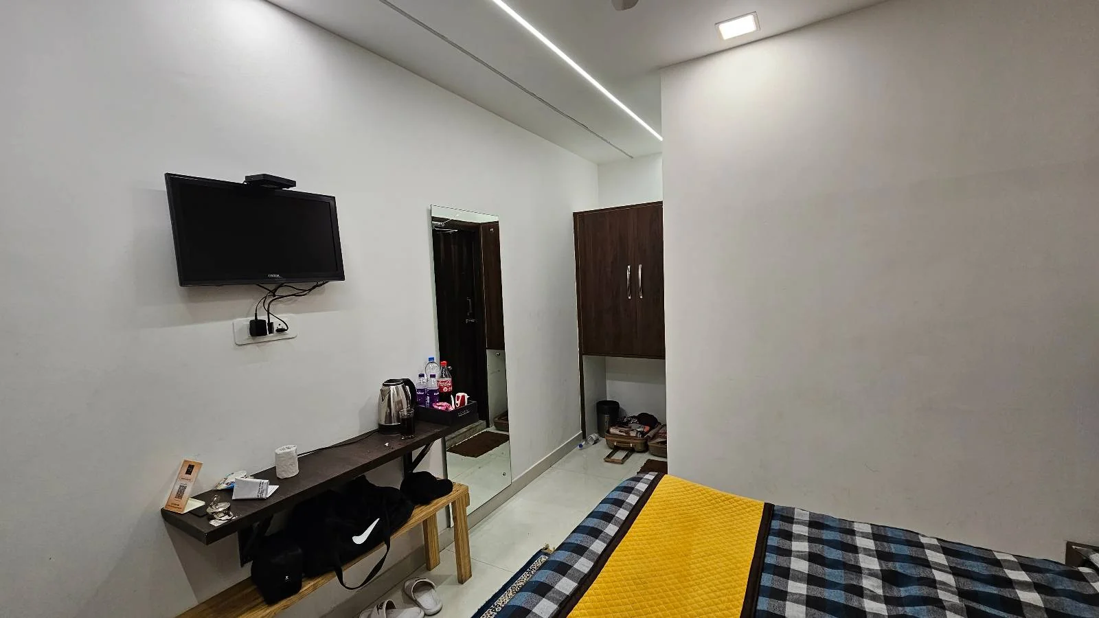 A small bedroom with a bed covered in a plaid and yellow blanket. There's a wall-mounted TV, a desk with items, a mirror, and a closet. The room has white walls and ceiling lighting.