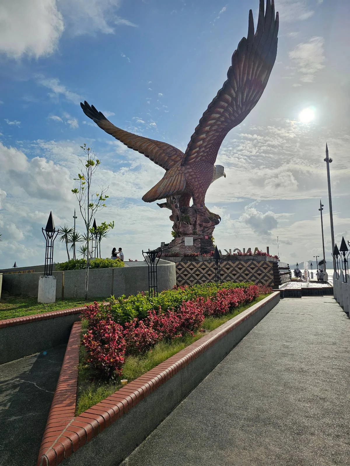 A large eagle statue with outstretched wings stands in a landscaped area near a waterfront under a bright, sunny sky with scattered clouds. Pathways and flowers are visible in the foreground.