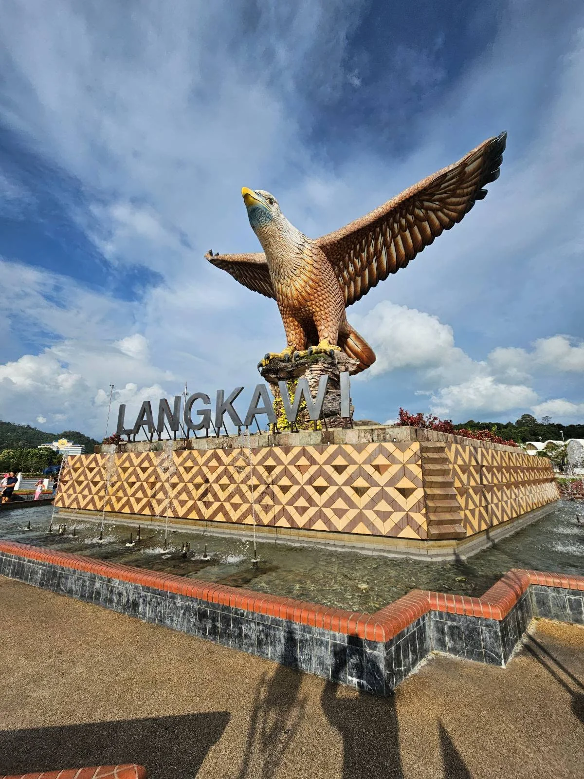 A large eagle statue stands on a decorated pedestal with the word "Langkawi" in big letters, under a blue sky with clouds. The area around the statue is paved and people’s shadows are visible.