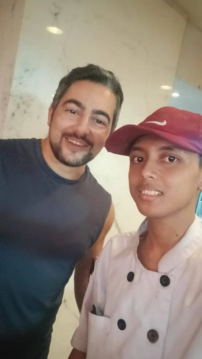 Two people smiling at the camera. The person on the left is wearing a sleeveless shirt, and the person on the right is dressed in a chef's outfit with a red cap. They are indoors with a light-colored wall in the background.