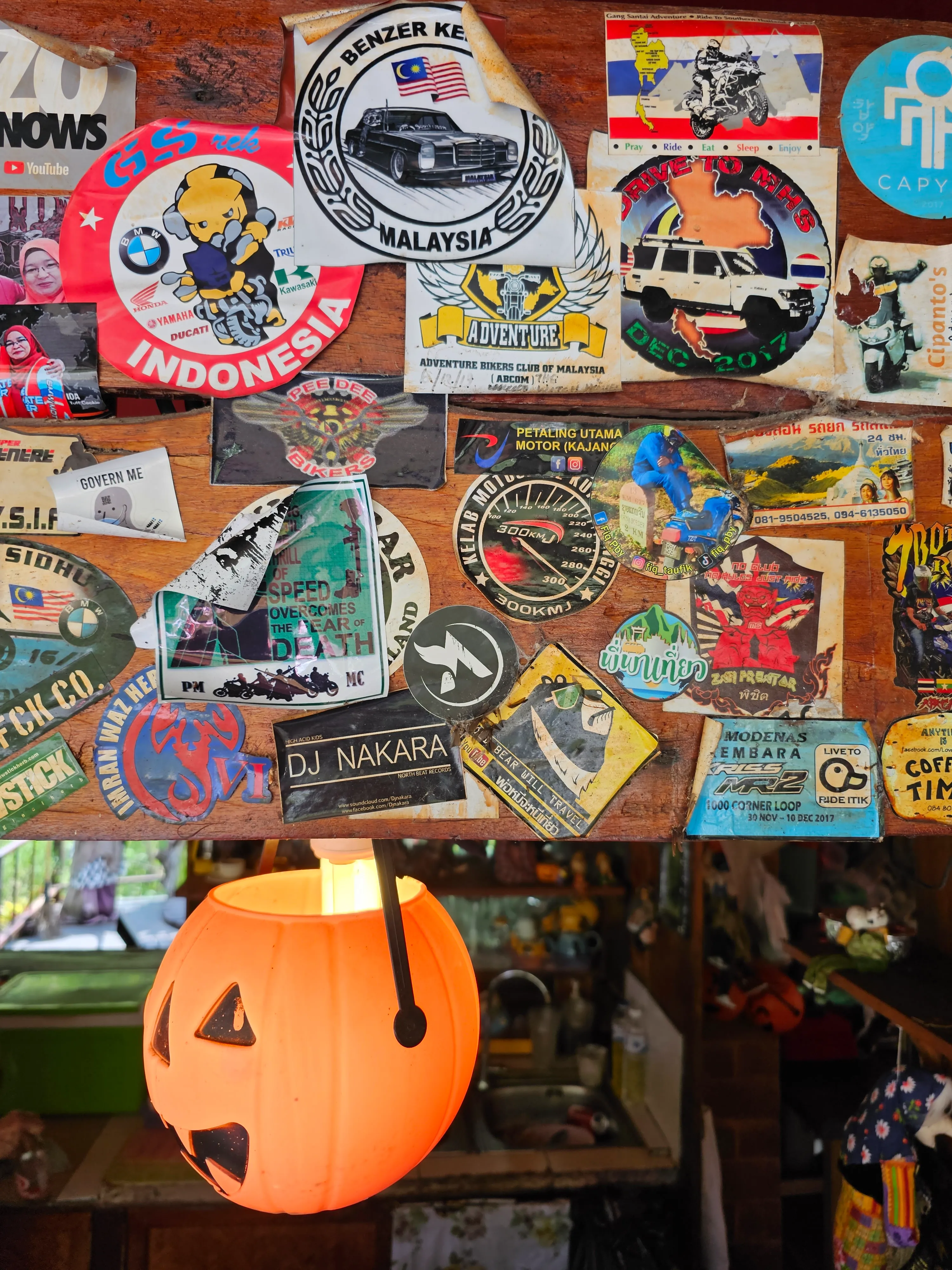 A collection of colorful travel and aviation-themed stickers covers a wooden surface. Below the stickers, a small orange paper lantern shaped like a jack-o’-lantern hangs, adding a festive touch.