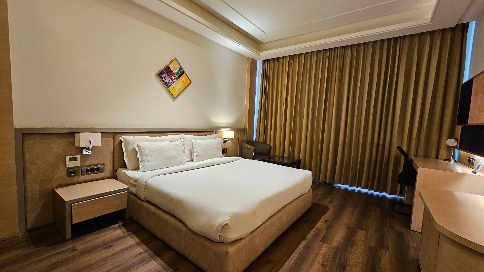 A modern hotel room with a large double bed, white linens, bedside tables, wall-mounted artwork, a desk with a chair, a TV, wooden flooring, and floor-to-ceiling curtains covering the windows.