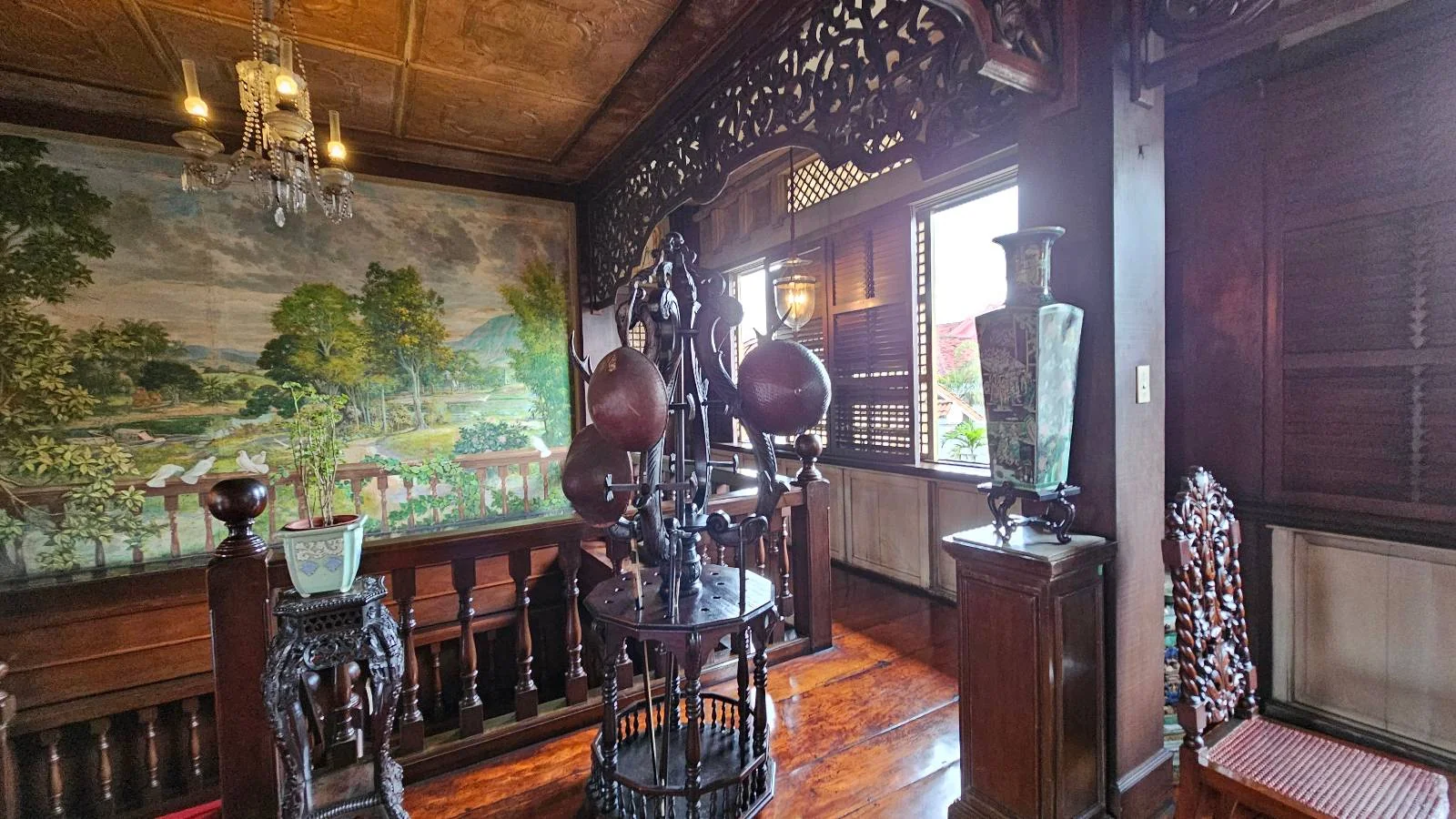 A room featuring intricate wooden furniture and decor, with a large mural of a landscape on the wall. A chandelier hangs from the ceiling, and sunlight streams through a window, illuminating ornate sculptures and vases.