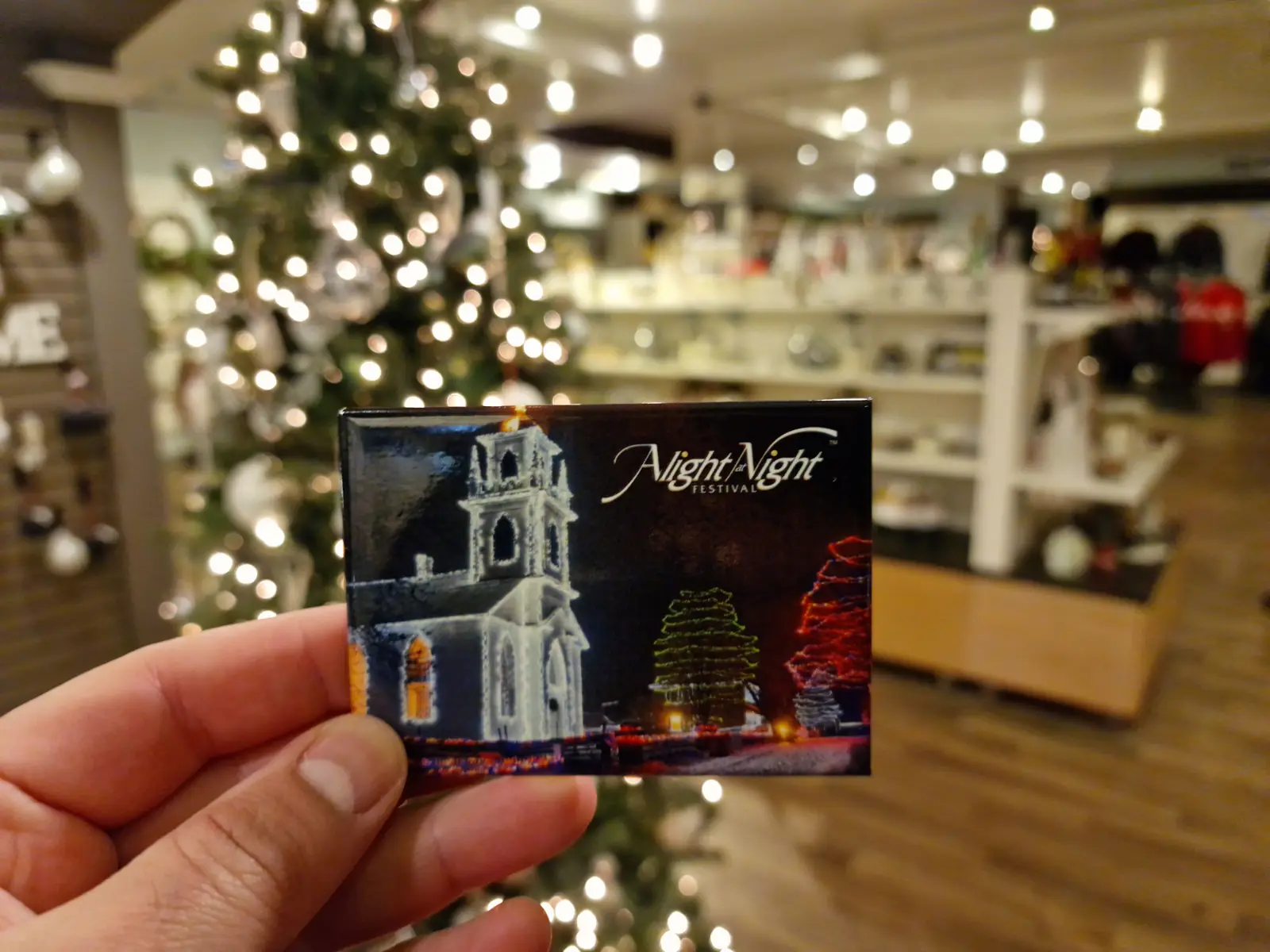 Hand holding a small postcard with a nighttime scene of a lit-up church and trees, inside a store decorated with Christmas lights and ornaments.