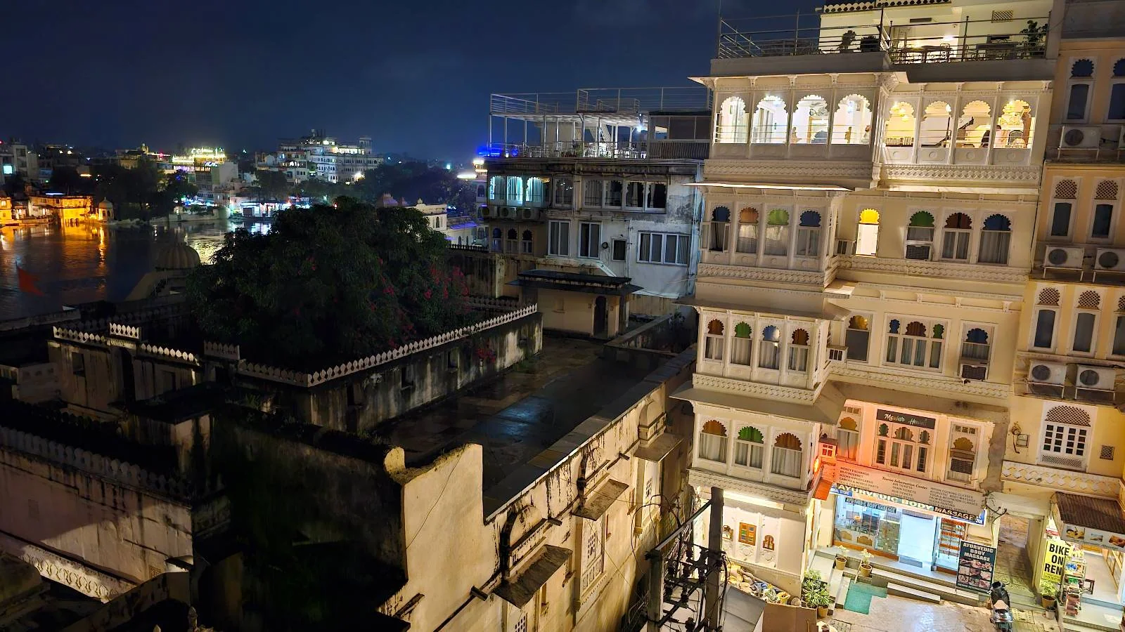 Nighttime cityscape showing brightly lit buildings with illuminated windows and balconies, overlooking a body of water. The scene includes various architectural styles, with city lights in the distance and a lively atmosphere below.