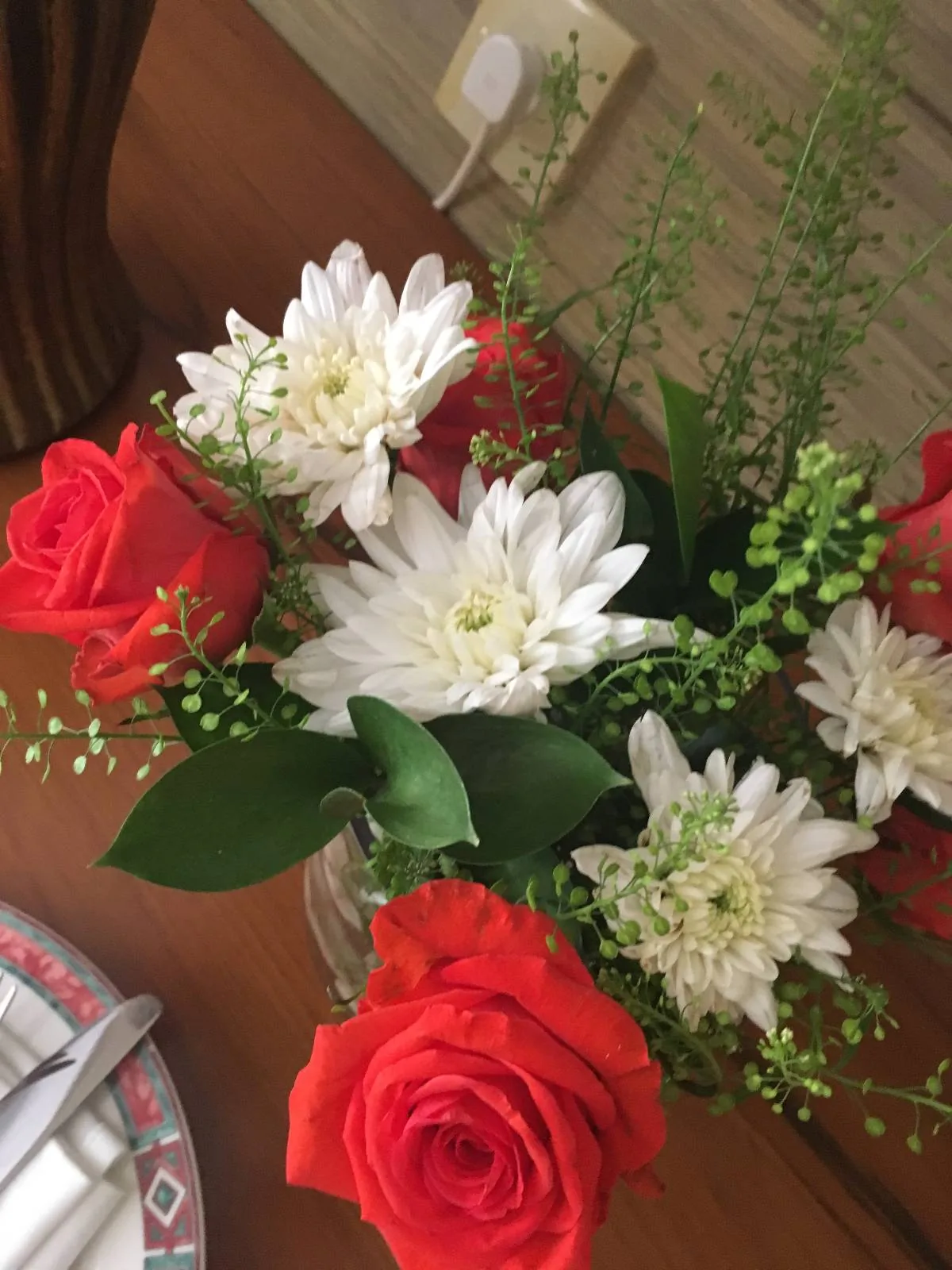 A bouquet featuring red roses, white chrysanthemums, and green foliage arranged in a clear glass vase. The flowers are placed on a wooden table near a decorative plate and an electrical outlet in the background.