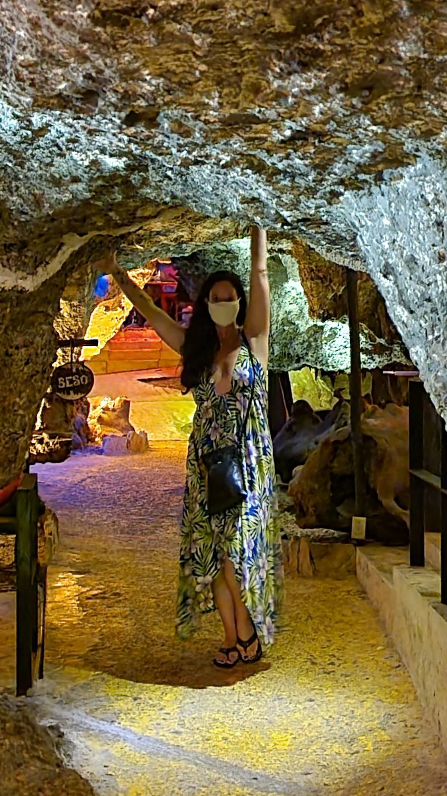 A person in a floral dress and face mask stands under a rocky archway in a cave, with colorful lights illuminating the background. They have one hand raised, touching the rock above. The setting appears to be a tourist attraction or natural site.