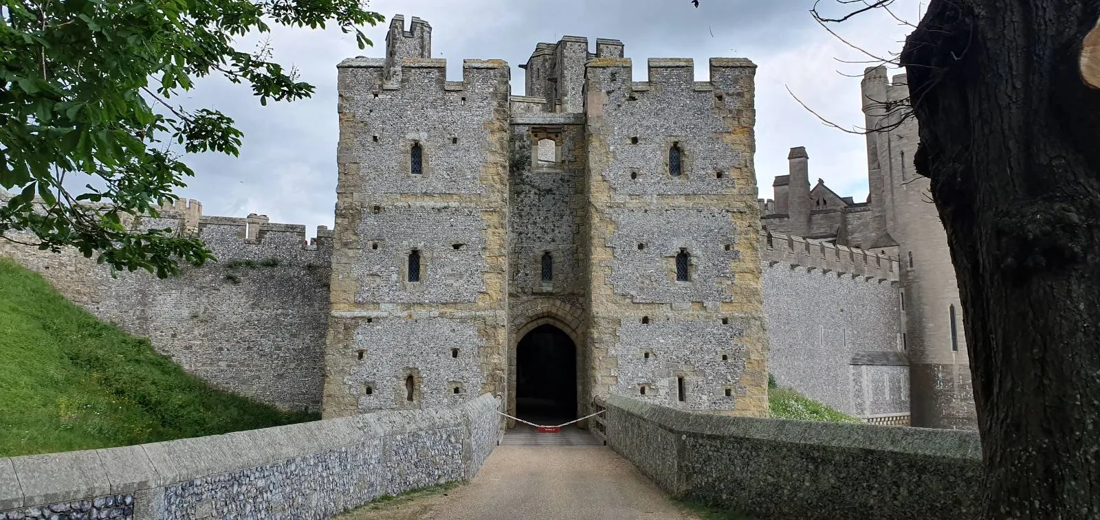 A stone castle with a tall, fortified entrance tower and turrets, surrounded by a grassy moat. The cloudy sky looms overhead, and a large tree is partially visible on the left. The path leading to the entrance is unpaved.