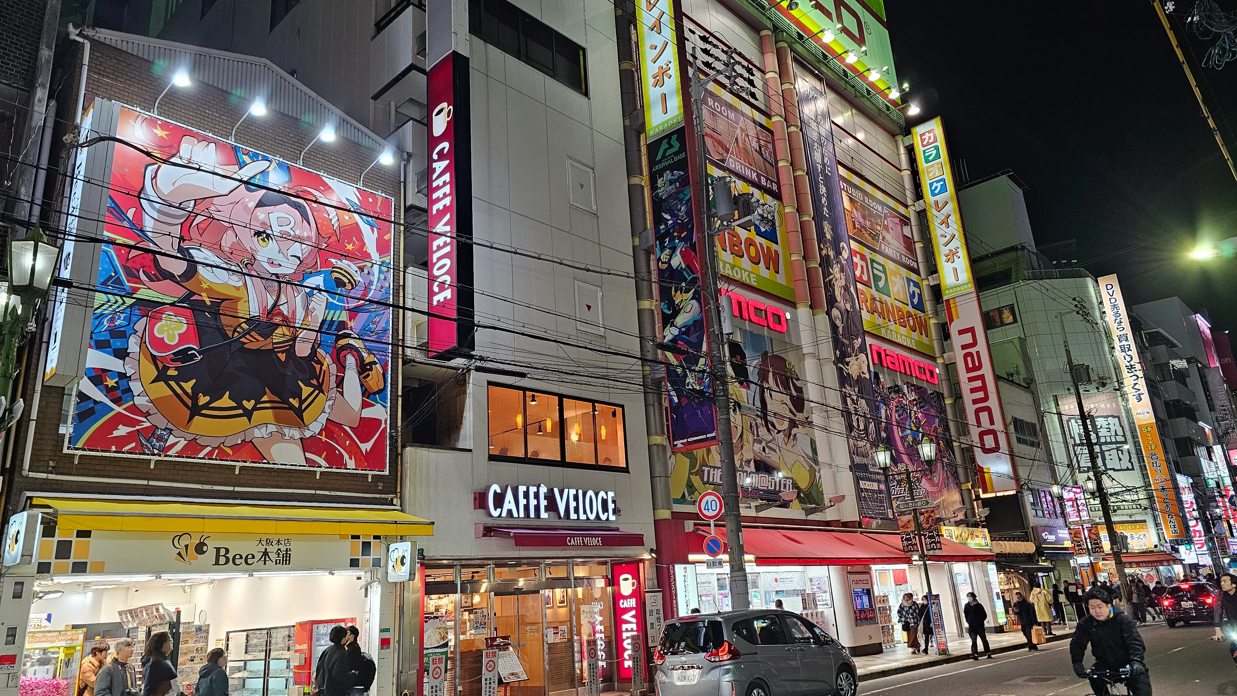 Colorful street scene at night in Akihabara, Tokyo, with bright anime billboards, illuminated shop signs, and people walking on the sidewalk in front of various stores and cafés.